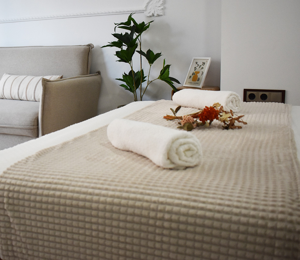 Vista parcial del interior: primer plano sobre cama con ropa de cama moderna blanca, colcha beige, toallas de baño enrollas blancas, ramo de flores. En el fondo el sofa gris claro con cojines beige con rayas marrones, molduras ornativas en la pared, planta.